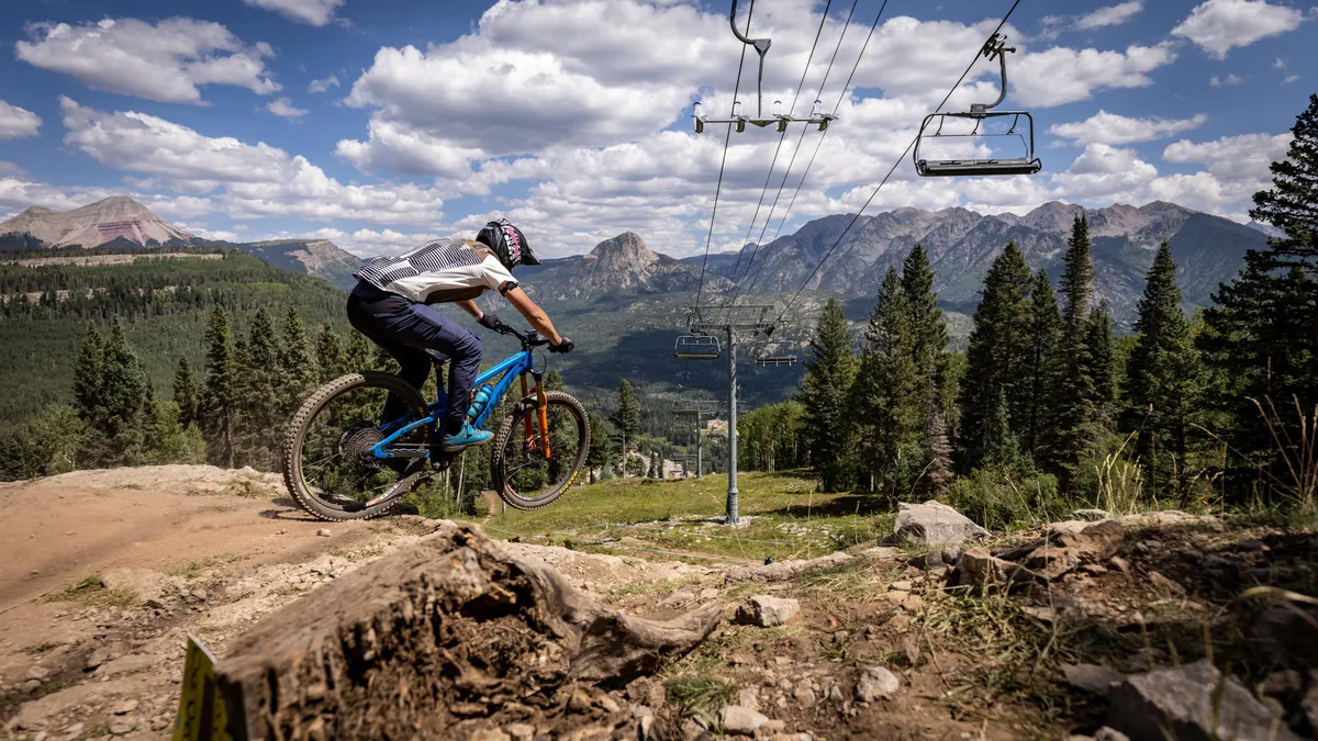 This exterior image captures a mountain biker riding along a scenic trail with the mountain range and ski lift in the background, surrounded by pine trees. The scene emphasizes outdoor adventure opportunities, highlighting both summer and winter activities available in the area.
