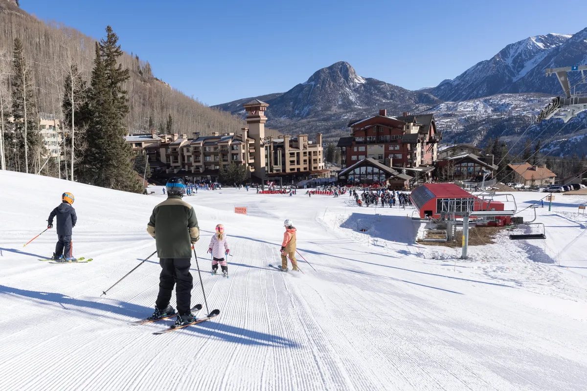 A dynamic outdoor scene showing skiers descending a snowy slope with the resort building in the background, mountains stretching into the distance, and a ski lift ready to whisk adventurers up the hill. The mood is adventurous and family-friendly, showcasing the excitement and scale of the local terrain.