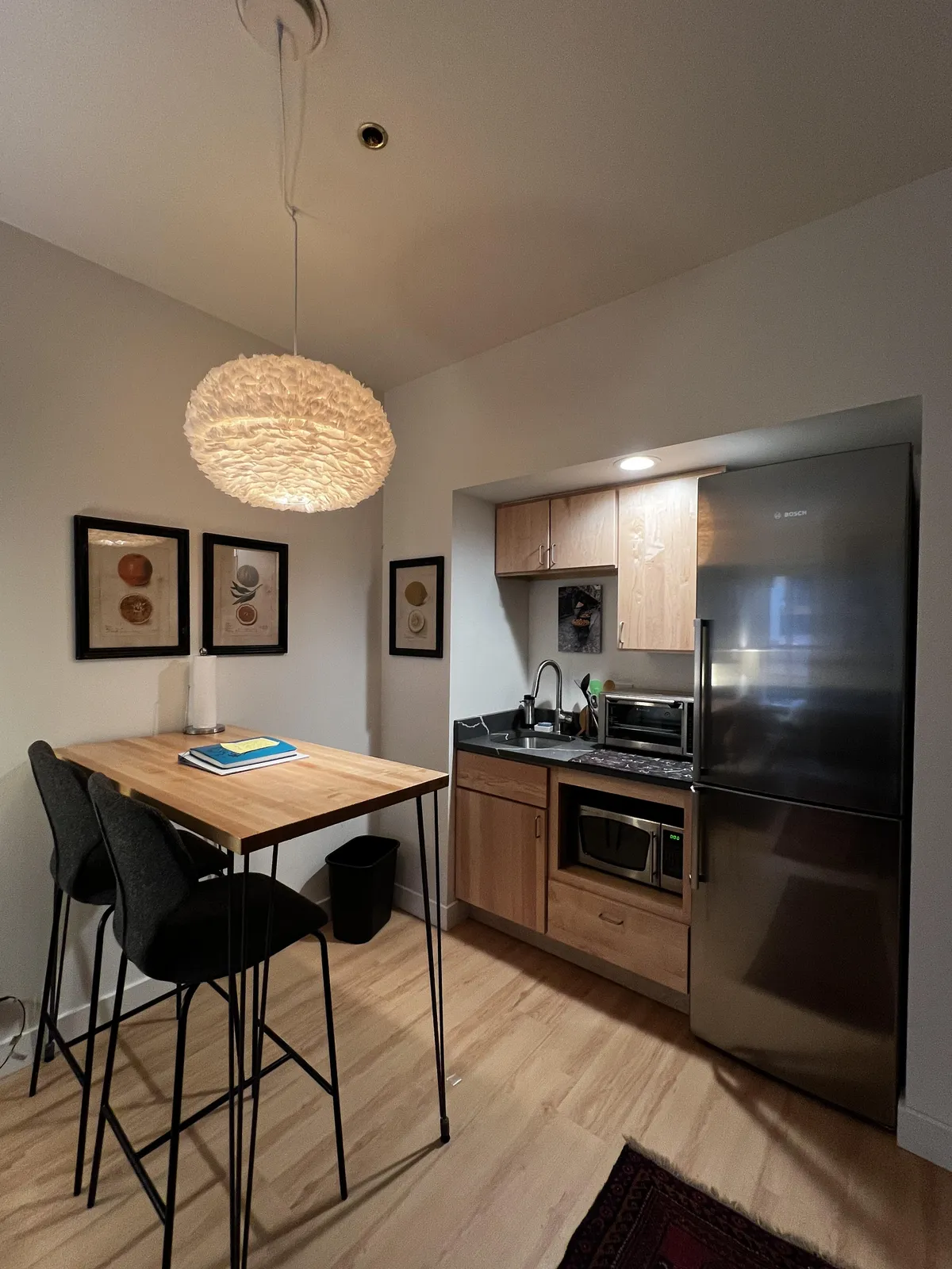 A contemporary interior scene featuring a wooden dining table, a modern hanging light fixture, and kitchen appliances in the background. Framed artwork adds personality, while the set-up suggests a space designed for both cooking and enjoying intimate meals.