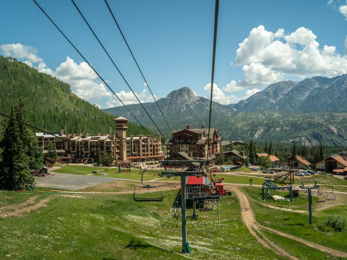 This image captures a ski lift ascending over lush green grass, with a breathtaking mountain range in the background, a rustic lodge, and clouds floating above. The vibrant setting speaks to both winter and summer activities, highlighting the allure of mountain adventures just steps from Kendall 341.