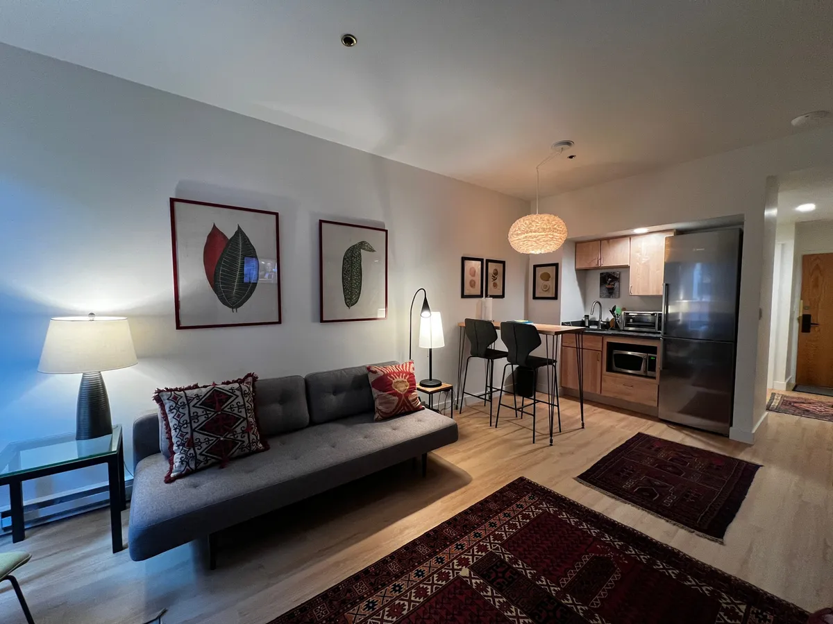 This image displays a stylish, well-lit interior with a sofa accentuated by decorative pillows, contemporary artwork on the walls, and a convenient kitchenette featuring bar stools at the counter and visible kitchen appliances. The comfortable setting suggests both relaxation and culinary activity, with the kitchen and casual dining space closely integrated.