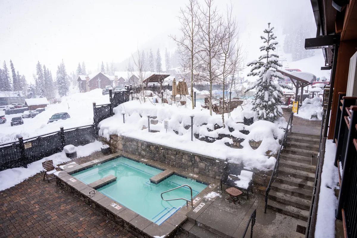 A serene outdoor hot tub sits nestled among snow-covered trees and a stone pathway, set against a tranquil winter landscape. The inviting steam from the hot tub contrasts with the wintry scene, creating a cozy and romantic atmosphere.