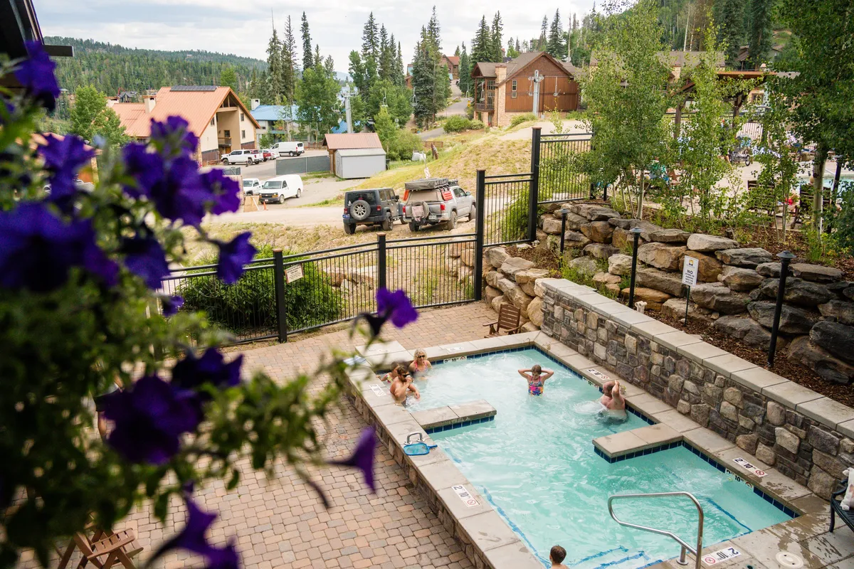 Set in a lush, vibrant landscape, this exterior shot highlights a hot tub surrounded by colorful flowers and greenery with majestic mountains in the background. The scene evokes relaxation, intimacy, and the beauty of summer evenings under the stars.