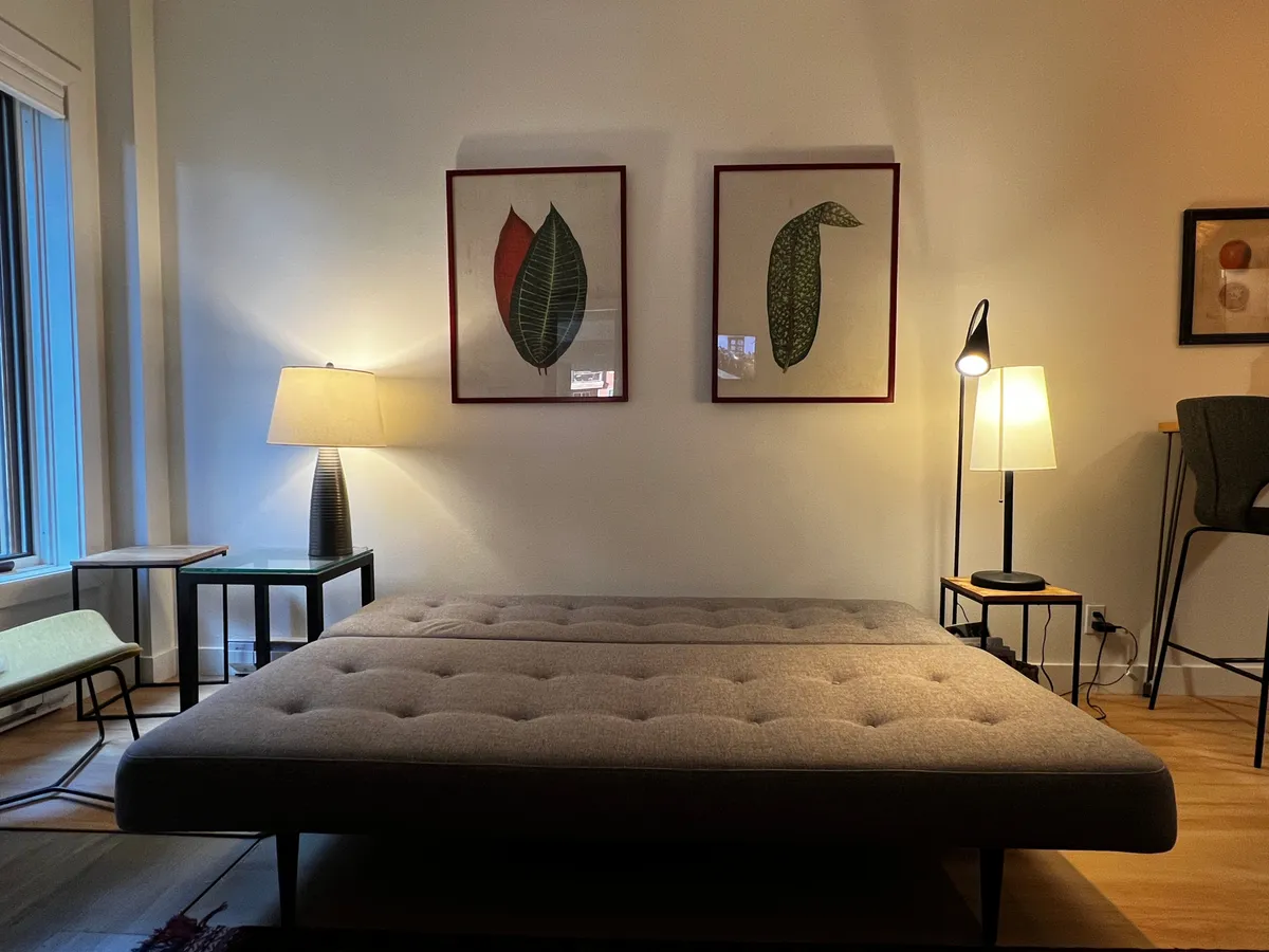 This image features a sofa bed positioned against a warmly decorated wall with tasteful mountain-inspired artwork and ambient table lamps casting a gentle glow. The cozy arrangement of furniture and accessories exudes comfort and intimacy, making it ideal for relaxation or quiet reading.