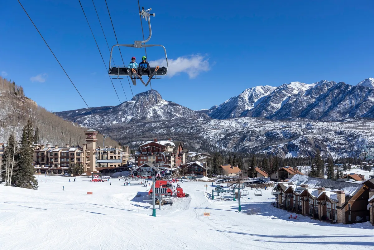 A wide view of snow-covered ski slopes, dotted with skiers and snowboarders, with the resort buildings and distant mountains in the backdrop. A ski lift traverses the scene, inviting guests to experience the excitement of the slopes or soak in the mountain scenery.