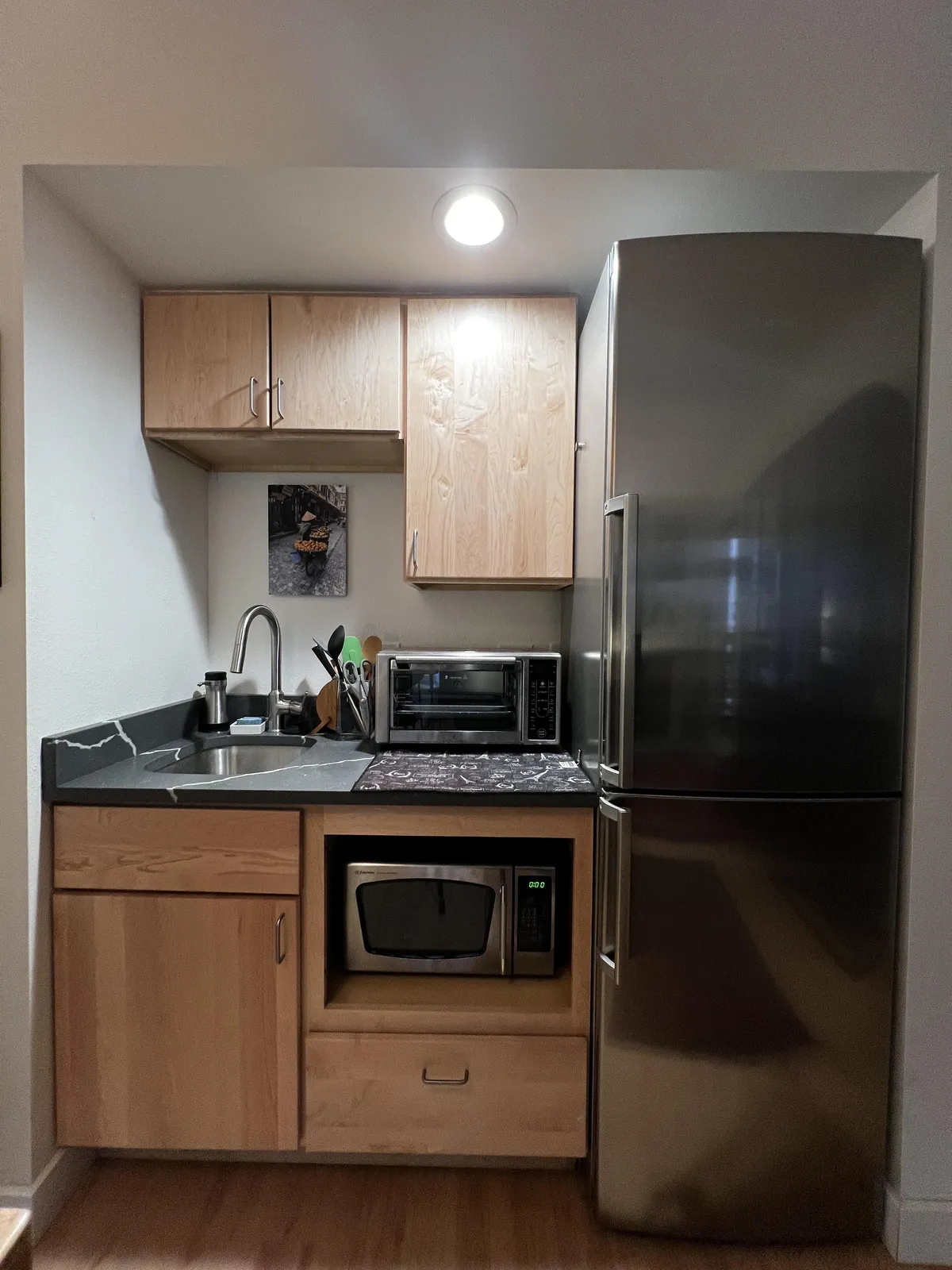 A functional kitchen interior focused on essential appliances: stainless steel refrigerator, microwave, sink, and wooden cabinets. The design is practical yet inviting, foregrounding the necessary amenities for self-catering and short stays.